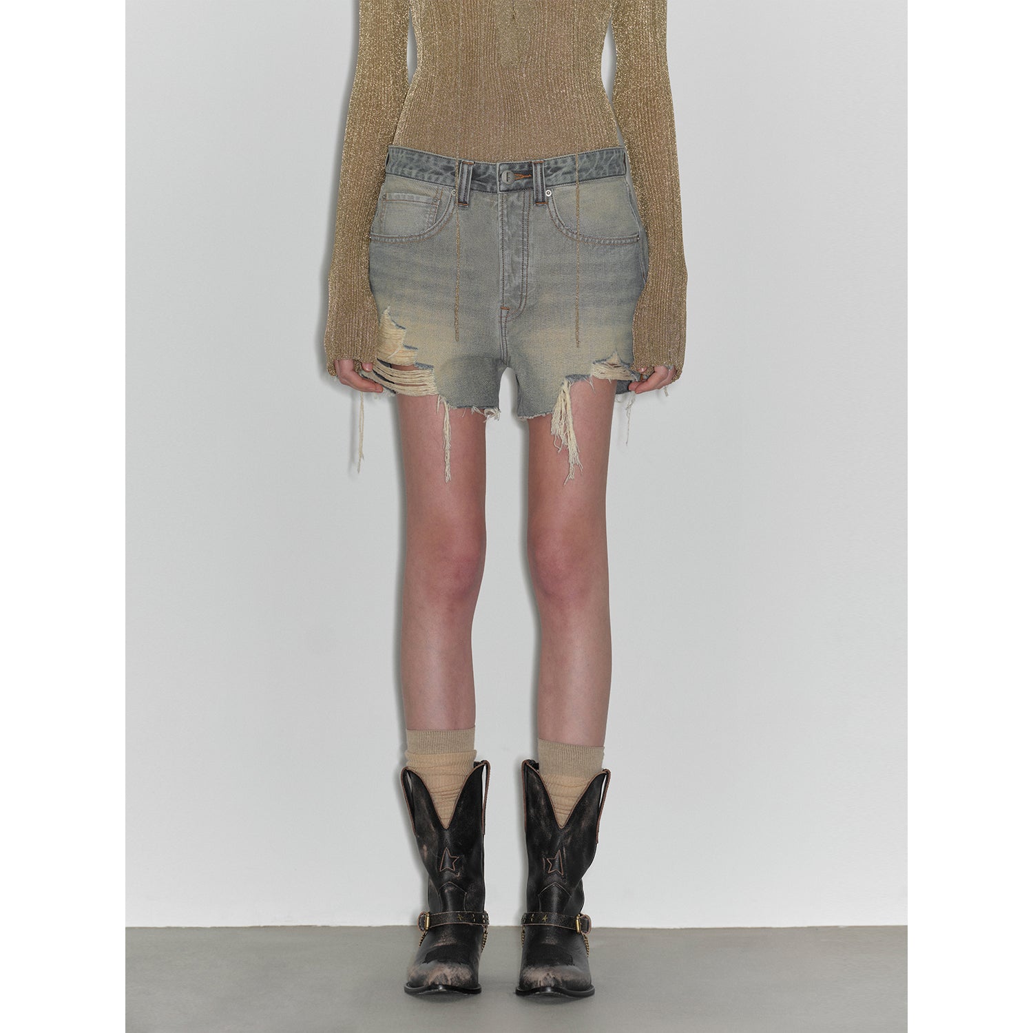 Person wearing distressed denim shorts and black cowboy boots on a plain background